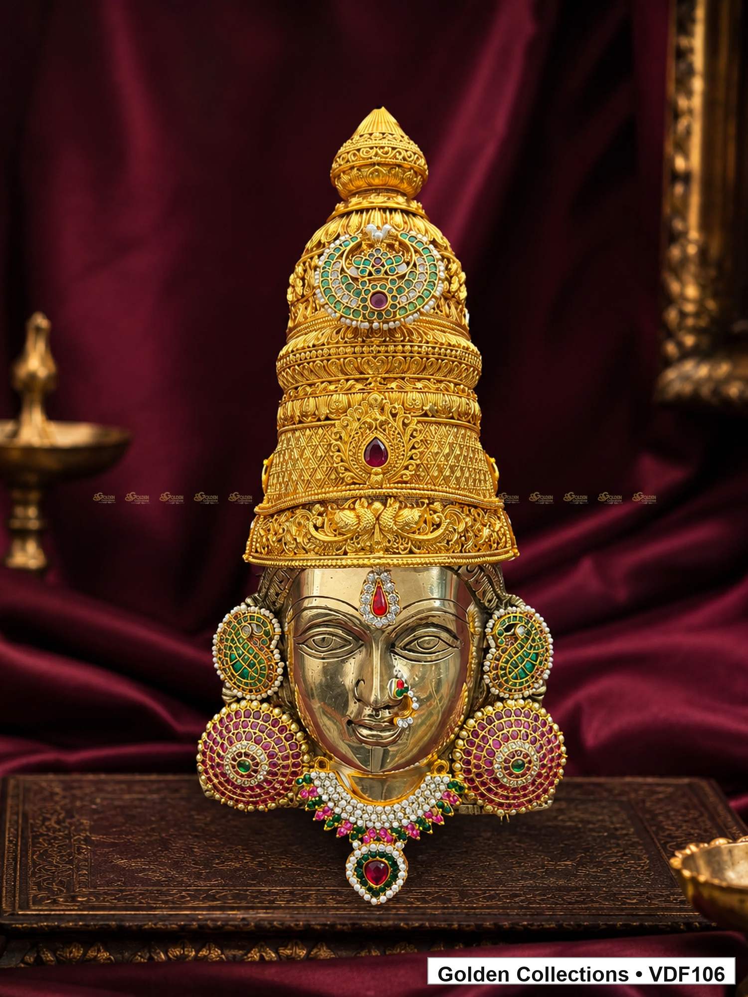 Fully Decorated Brass Varalakshmi Face with Nakshi Crown and Green Jadau Earrings displayed against a burgundy devotional backdrop