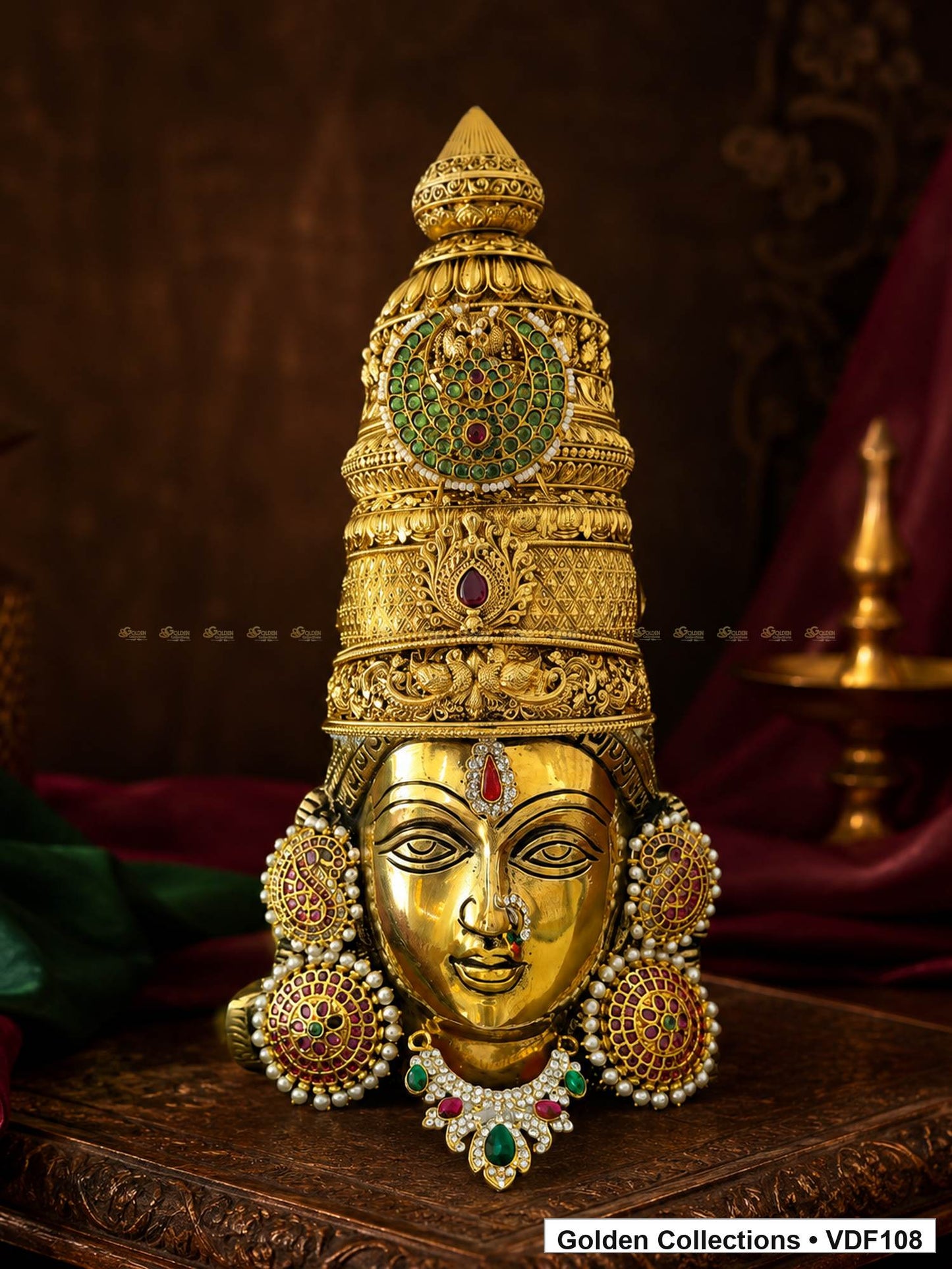 Fully Decorated Brass Varalakshmi Face with Nakshi Crown and Green Teardrop AD Necklace displayed against a burgundy devotional backdrop
