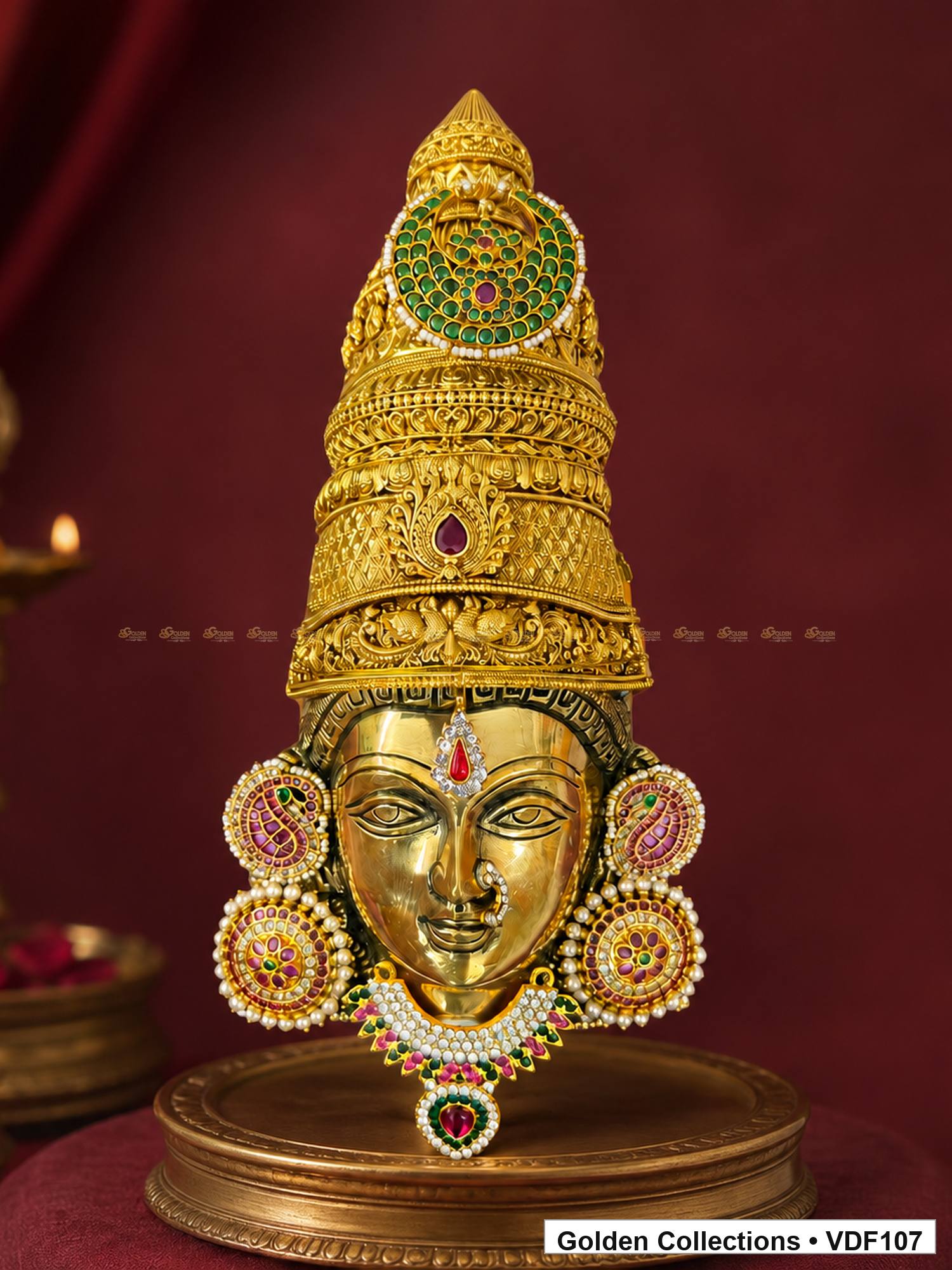 Fully Decorated Brass Varalakshmi Face with Nakshi Crown and Pink Jadau Earrings displayed against a burgundy devotional backdrop